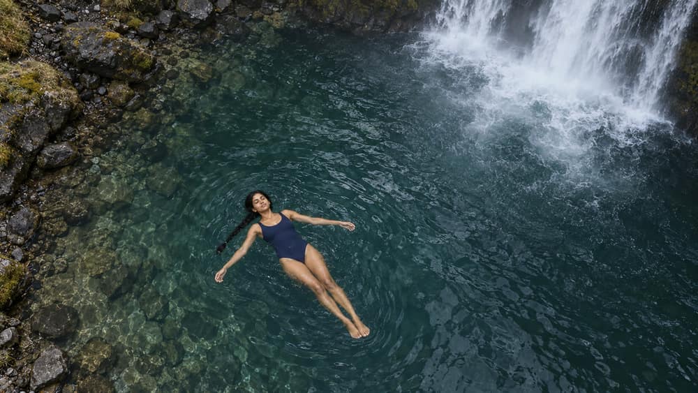 Panel 08, Floating: Elevated wide view of Nisha lying face-up in the pool, with the waterfall entering the top of frame.