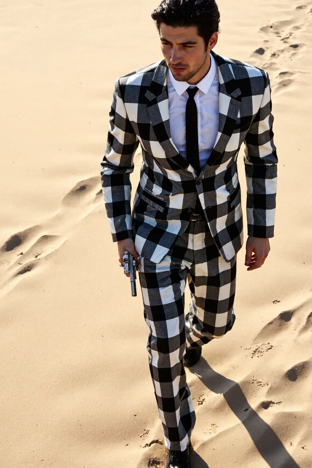 A man in a checked suit carries a revolver as he walks across sunlit sand.