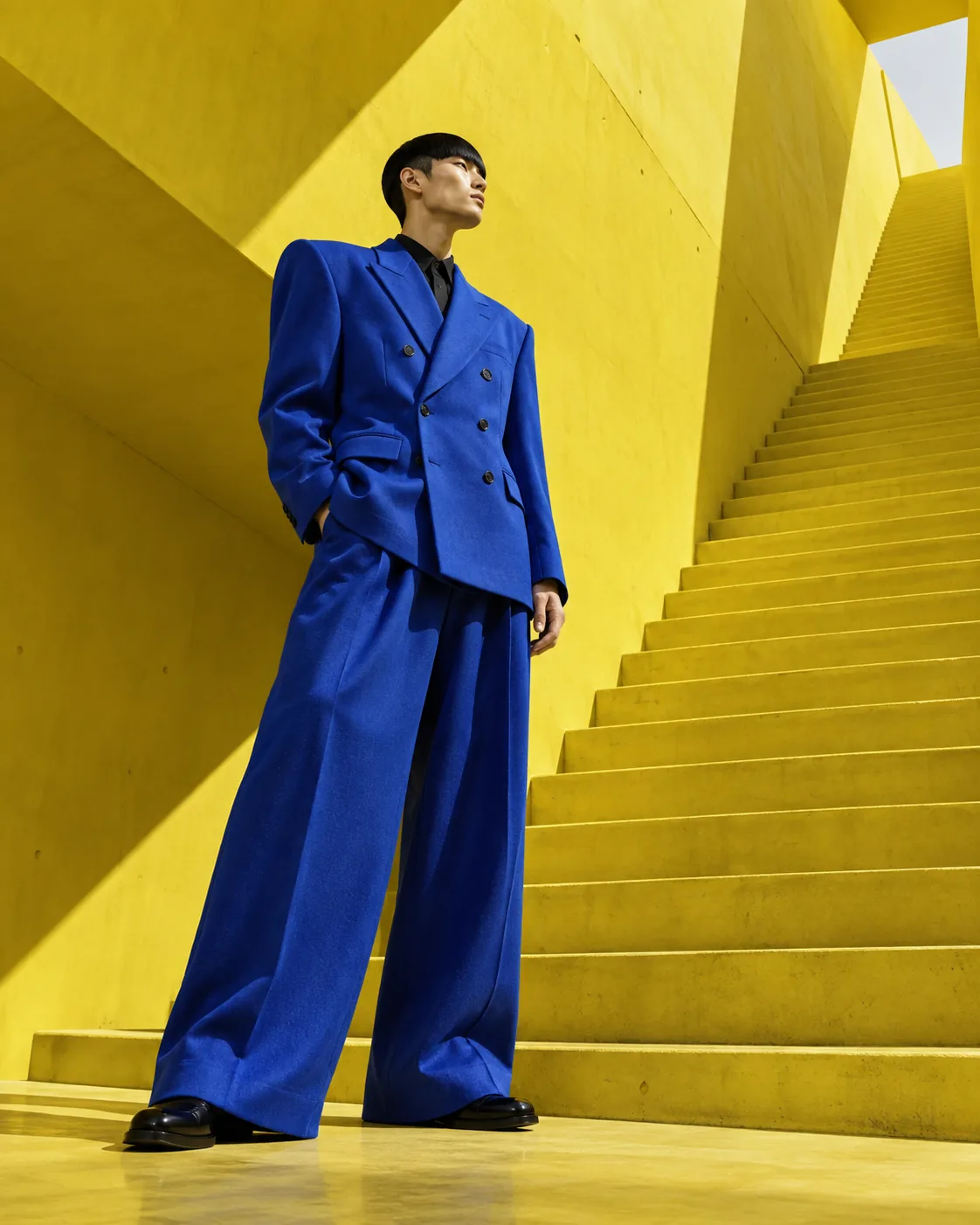 A man in a cobalt-blue double-breasted suit stands on a bright yellow staircase.